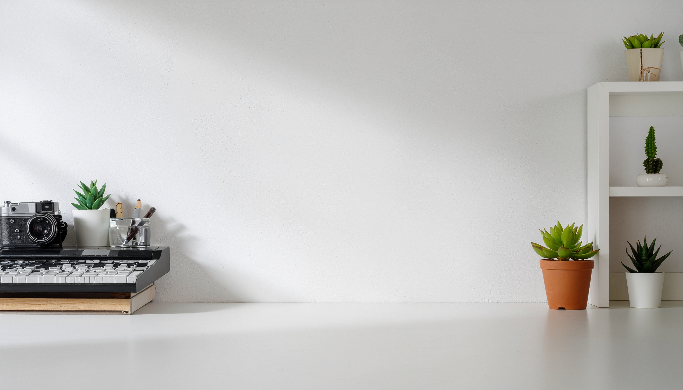 Espaço minimalista com câmera vintage e teclado sobre mesa branca, ao lado de suculentas em vasos e estante branca ao fundo, iluminado por luz suave e natural, criando uma atmosfera serena e organizada.